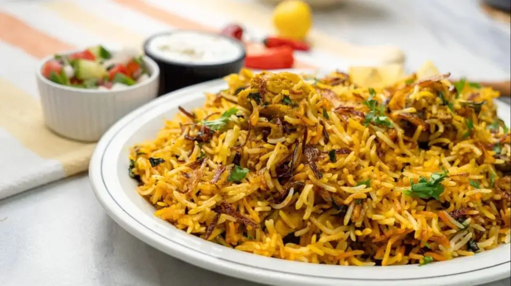 A plate of flavorful chicken biryani garnished with fried onions and fresh coriander, served with bowls of salad and raita on the side.