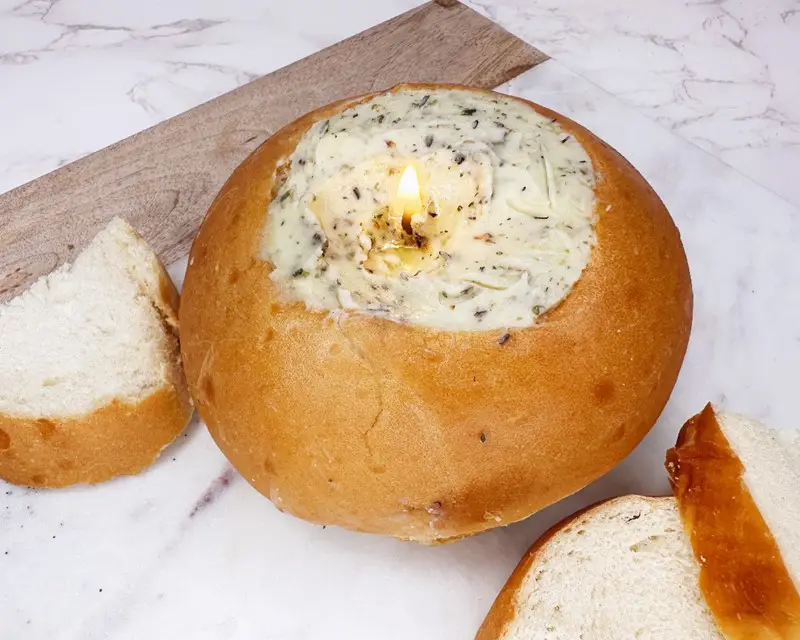 A hand holds a piece of bread, dipping it into a lit, green-hued herbed butter candle, surrounded by a scored loaf of bread.