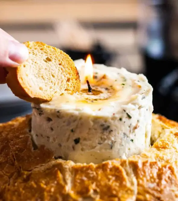 A hand holding a piece of light brown bread dips it into a melted, orange-brown candle-like substance with a burning wick, set within a larger, round loaf of bread.