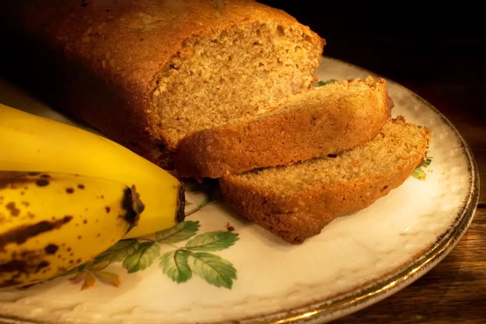 A loaf of banana bread with two slices cut, resting on a decorative plate next to ripe bananas, against a dark background.