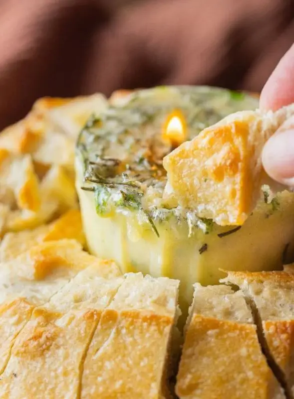 An overhead shot of a lit butter candle, made of herbed butter, melting in the center of a round bread bowl on a white marble surface with sliced bread beside it.