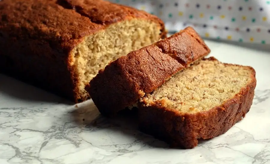 A sliced loaf of banana bread with walnuts on a wooden cutting board, with fresh bananas in the background.